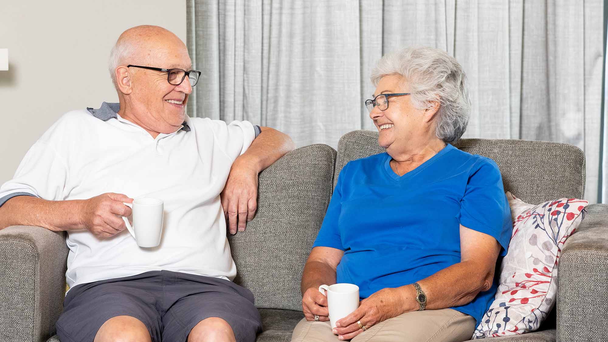 An older man and woman sitting on a couch and having a hot drink