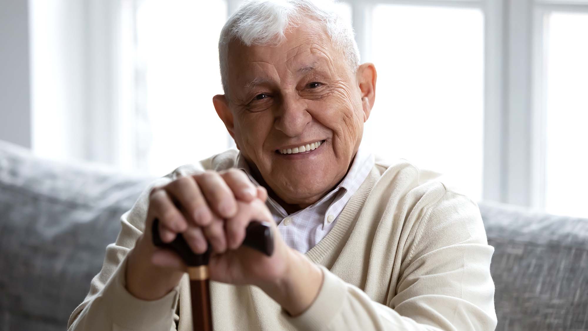 An older man smiling and sitting on a couch with his hands on a walking stick