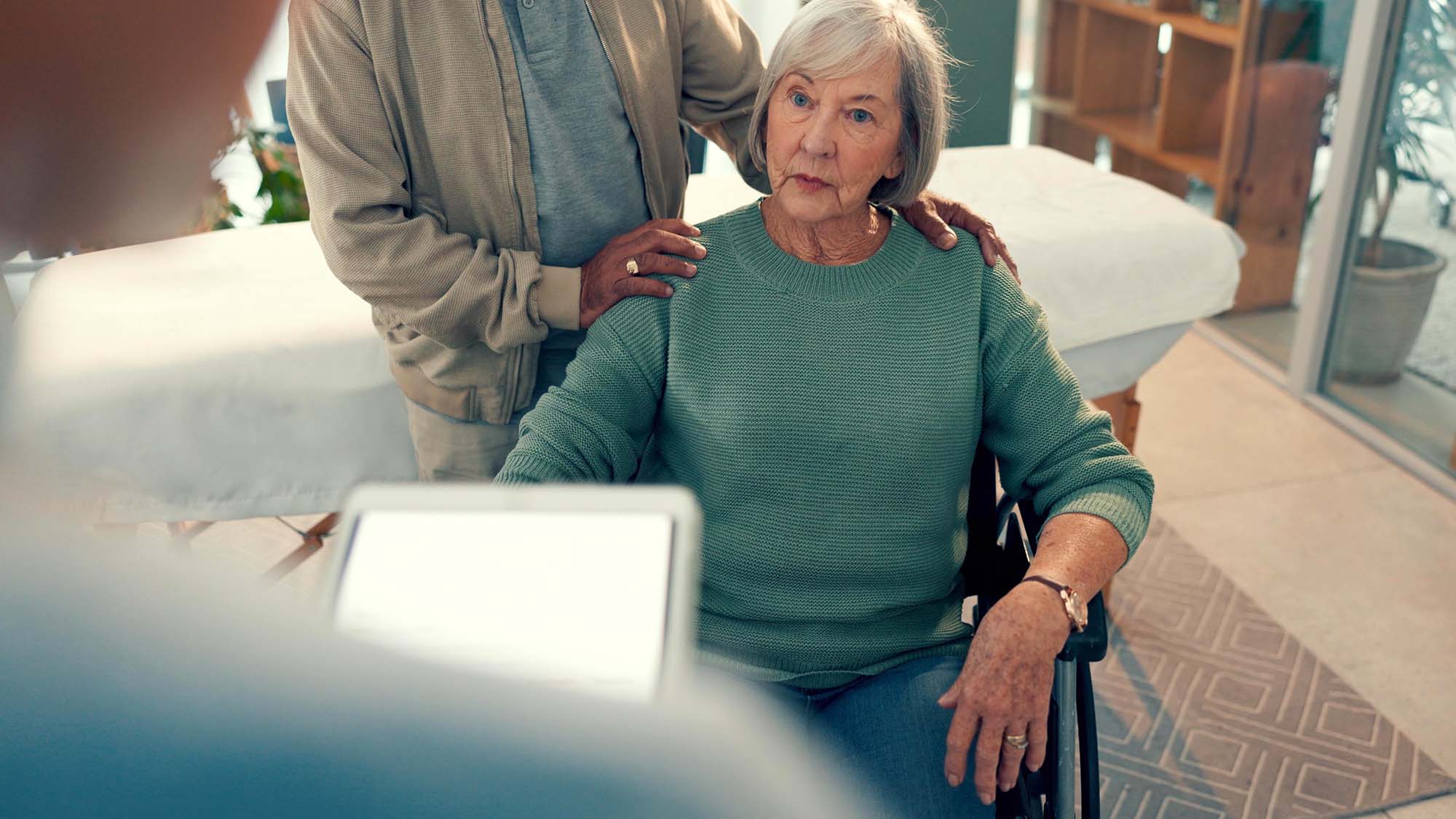 An older woman in a wheelchair listening to a doctor with someone standing behind her with their hands on her shoulders