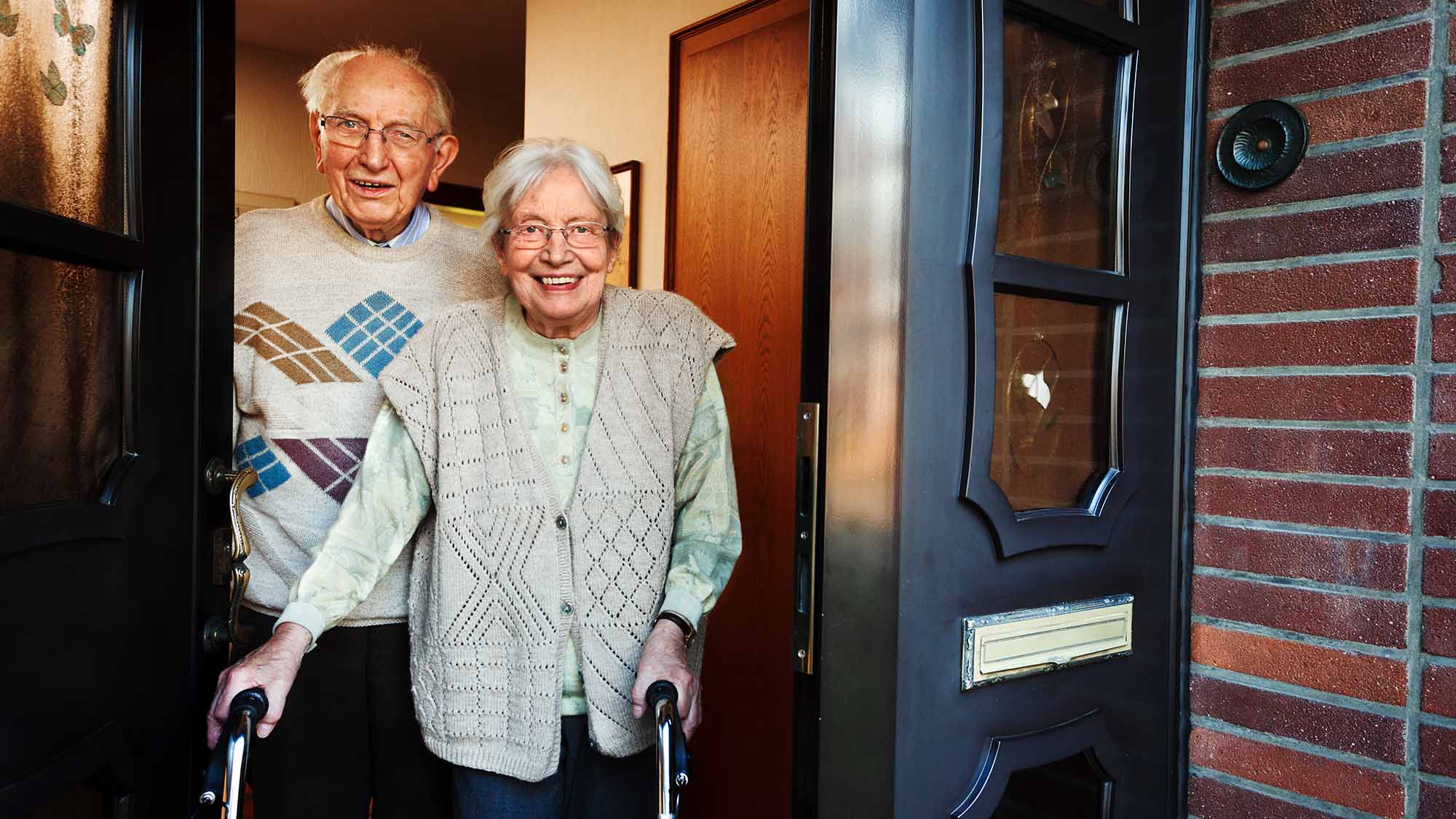 An older woman holding a walking aid and an older man standing in a doorway 