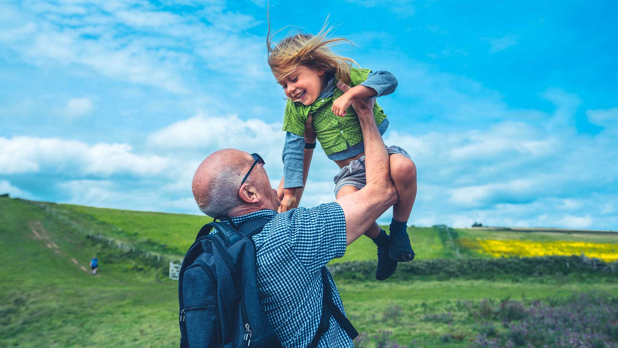 An older man lifting a young child in the air while being outside in a field